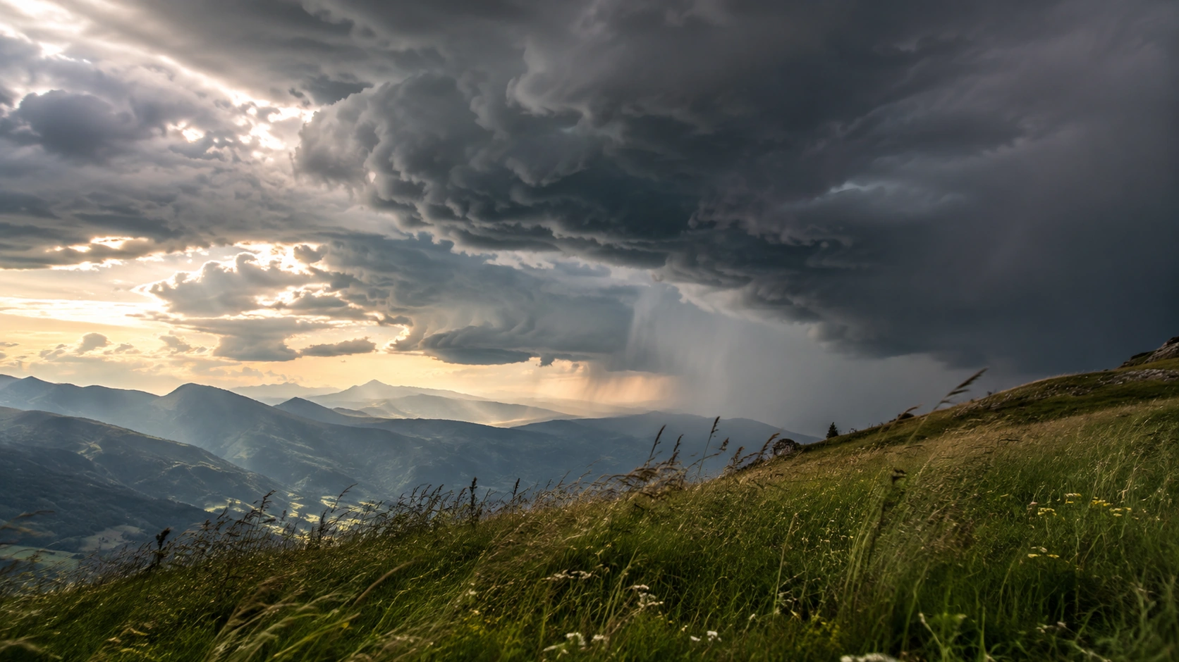 Berglandschaft unter markanten dunklen Gewitterwolken mit sichtbarem Niederschlagsvorhang in der Ferne.